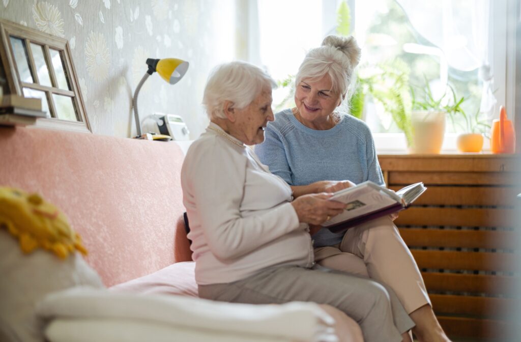 Older parent and their adult child sit together on a couch, smiling and looking at a photo album in a warmly lit room.