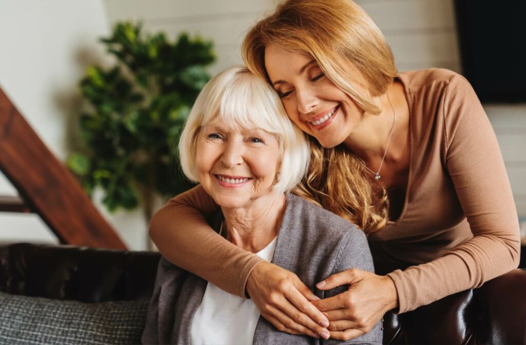 A smiling adult child hugging their senior parent's shoulders from behind while they sit on the couch.