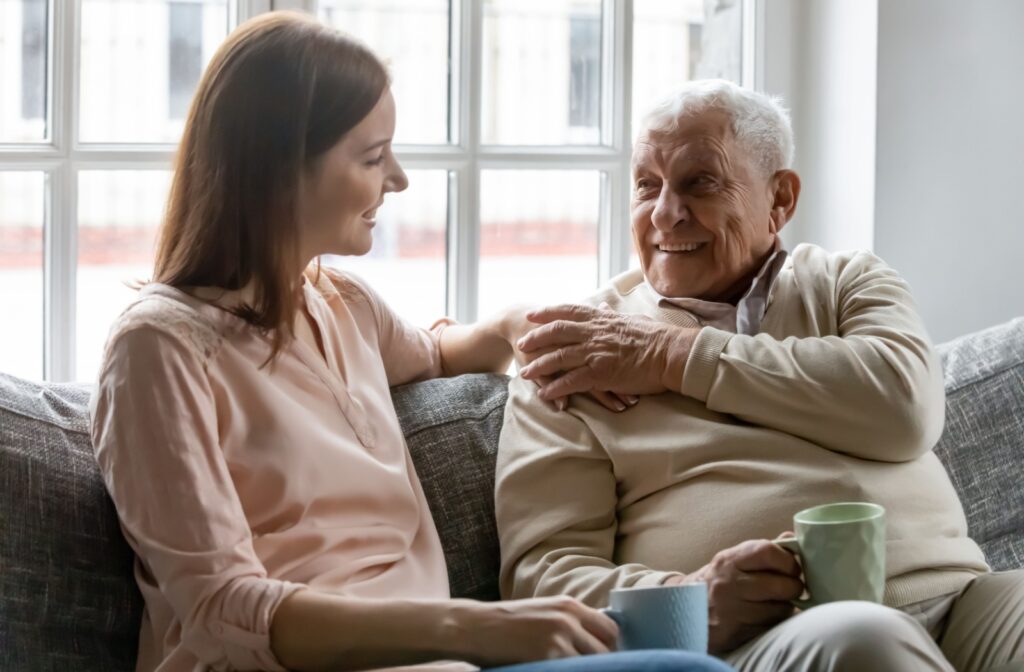 Young woman and older adult man sitting on a couch with mugs talking. Woman has hand on man's shoulder, with man's hand on top.