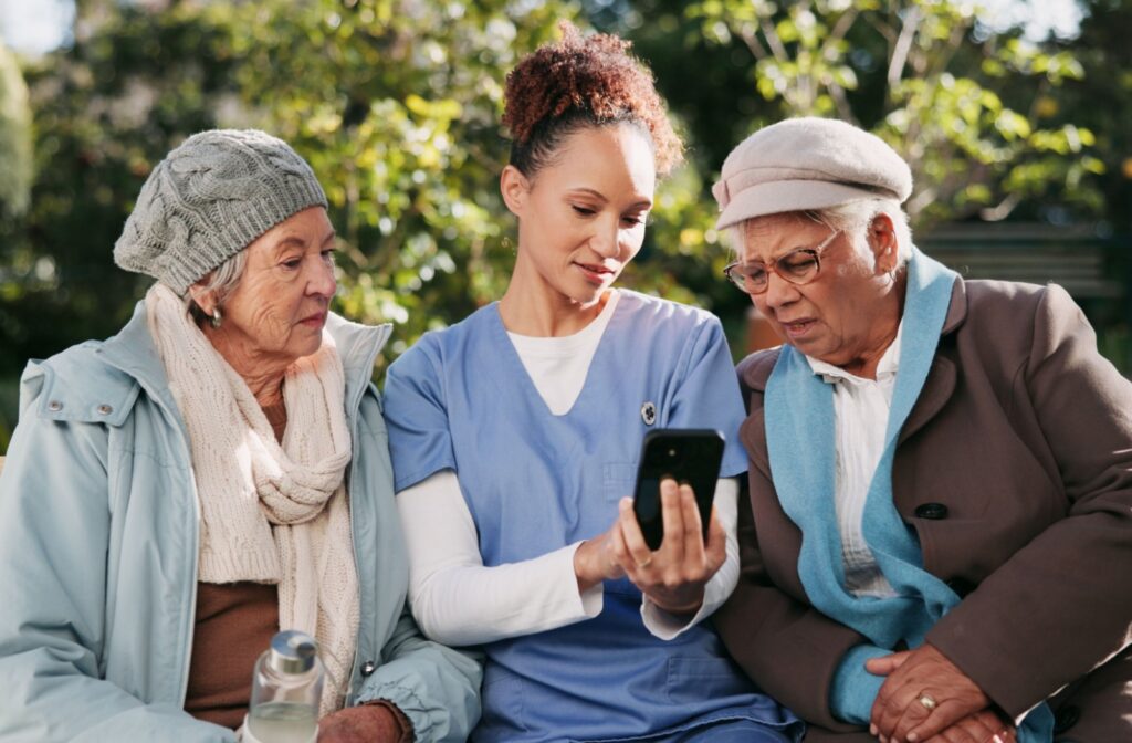 A nurse sitting outdoors between two seniors shows them something on their phone while providing help in assisted living
