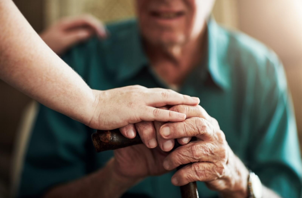 An older adult rests their hands on top of their cane and prepares to stand while a caregiver gently touches their fingers to show support