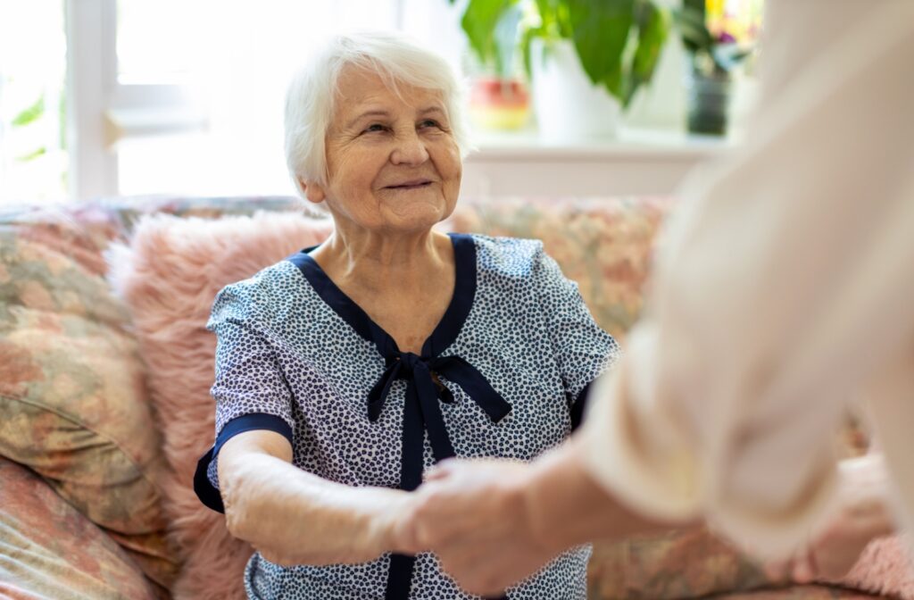 An older adult smiles while an out-of-frame caregiver grabs their hands to help them stand up from the couch in their living room in assisted living