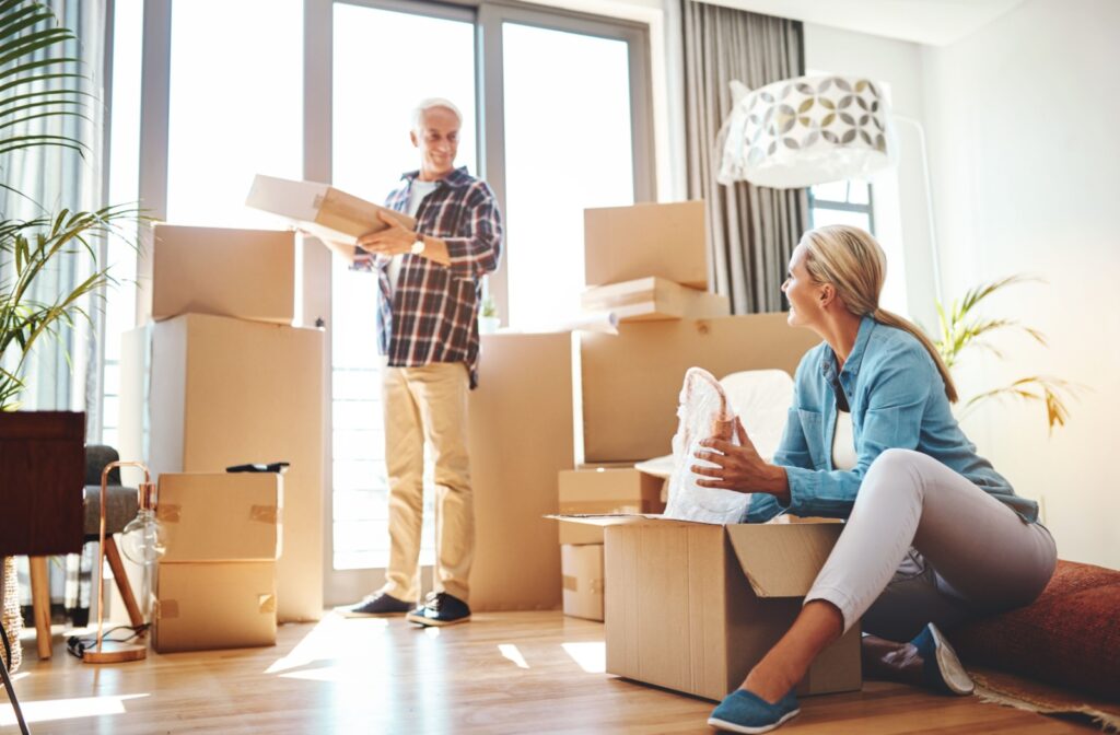 An older adult stands in front of a window in their packed-up home while lifting a box and smiling at their seated spouse on the floor
