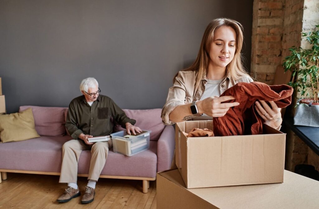 A young adult carefully folds a shirt and packs it in a box to help pack while their older parent sits on a couch and sorts through documents in a clear box